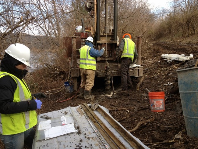 Workers in yellow vests use equipment to bore into the soil.